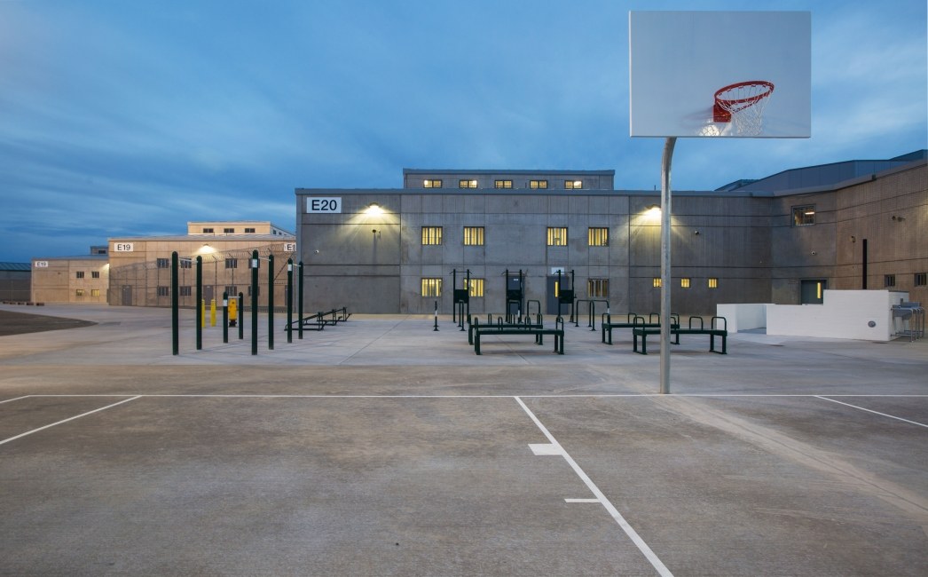 Dusk view across the exercise yard at Mule Creek State Prison, with the two-story concrete housing units E19 and E20 lit from within, outdoor exercise equipment and a basketball court in the foreground.